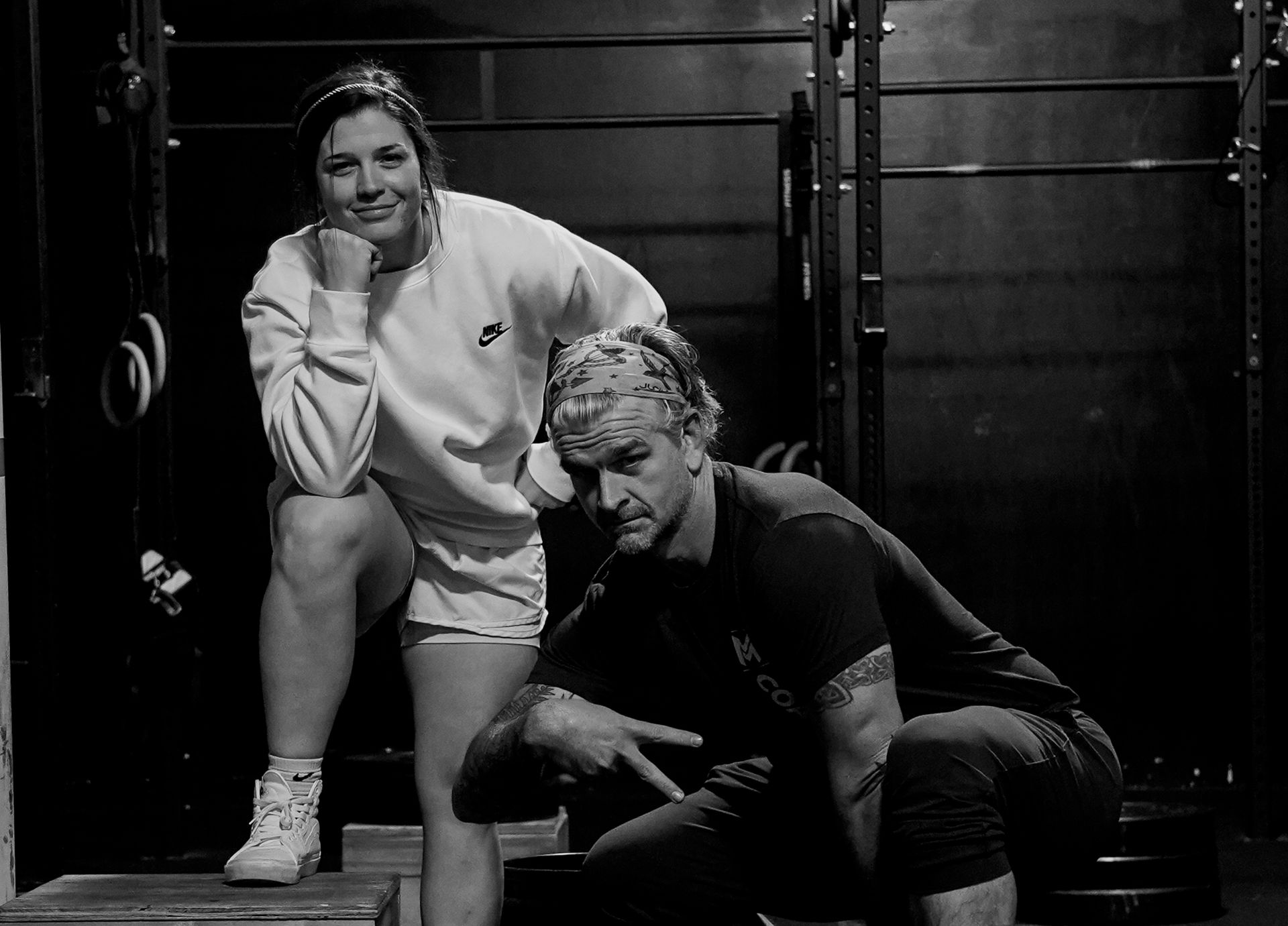 Two people posing in a gym, one seated and one crouching, both in athletic wear.
