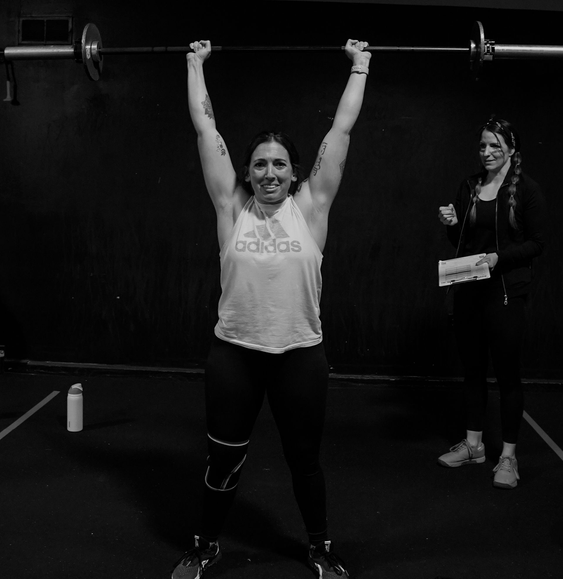 Woman lifting a barbell overhead in a gym while another person watches nearby