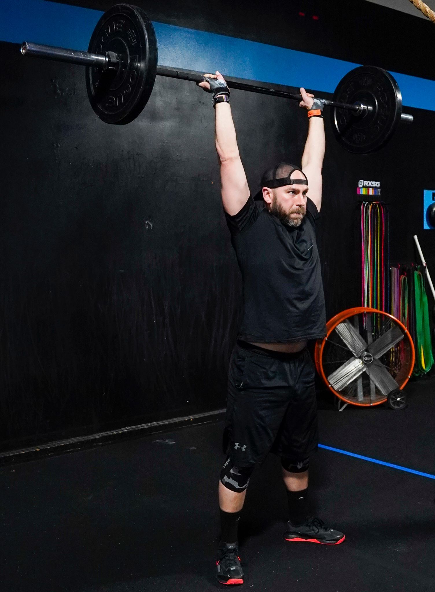 Man lifting a barbell overhead in a dark gym, wearing black workout clothes and knee sleeves