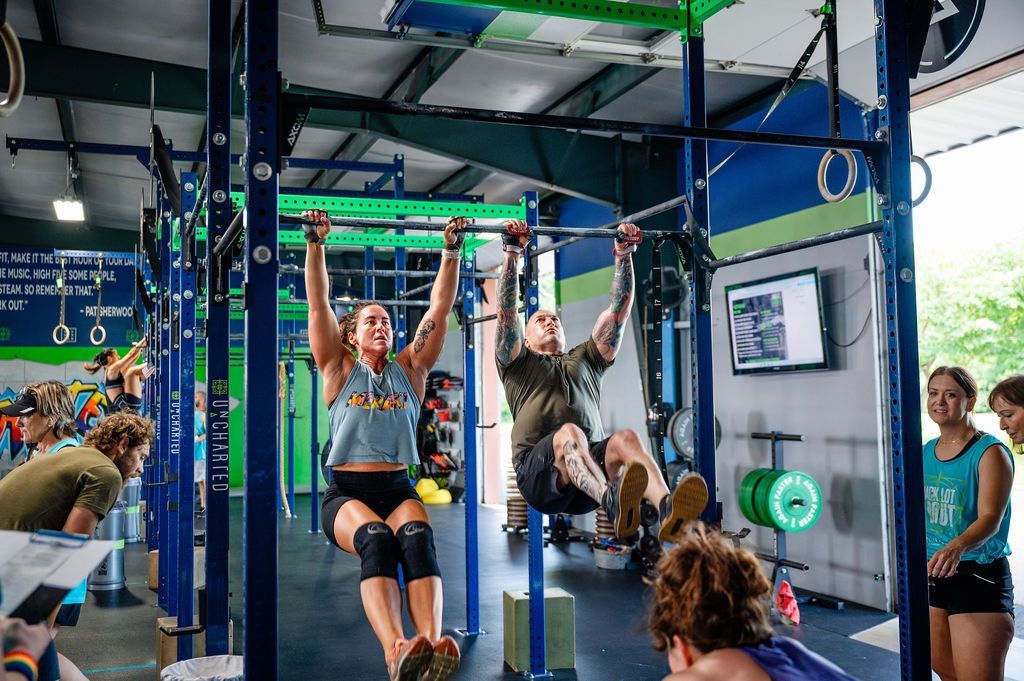 People exercising on hanging straps in a blue gym with neon rings and a wall-mounted screen.