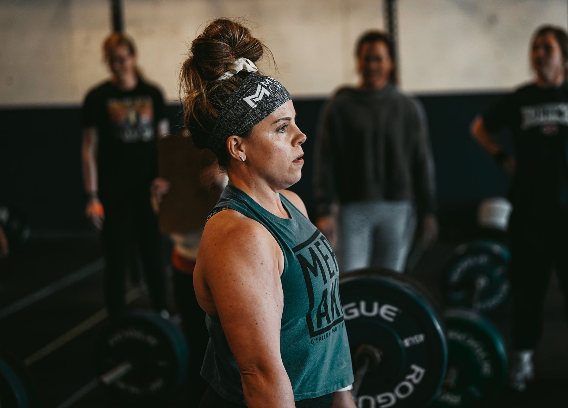 Woman in green workout gear preparing to lift a barbell in a gym, with teammates in the background
