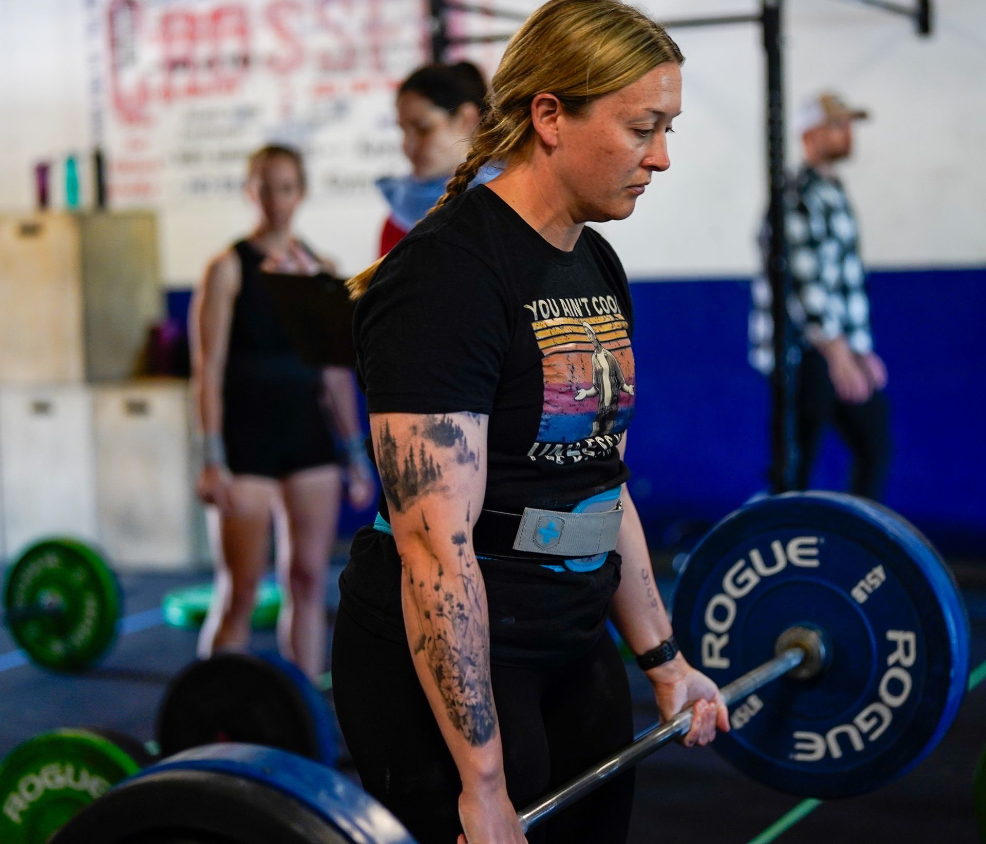 Person lifting a barbell in a gym, with bumper plates and other people blurred in the background.