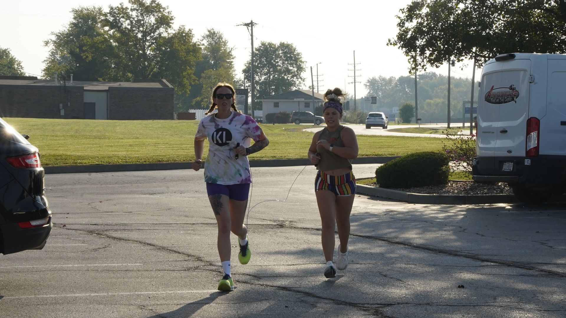 Two runners in athletic gear jogging on a paved path near parked vehicles in a sunny lot