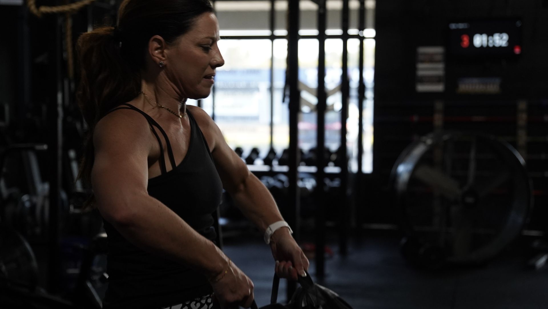 Woman resting with a gym bag in a dimly lit gym, near workout equipment and a digital clock.