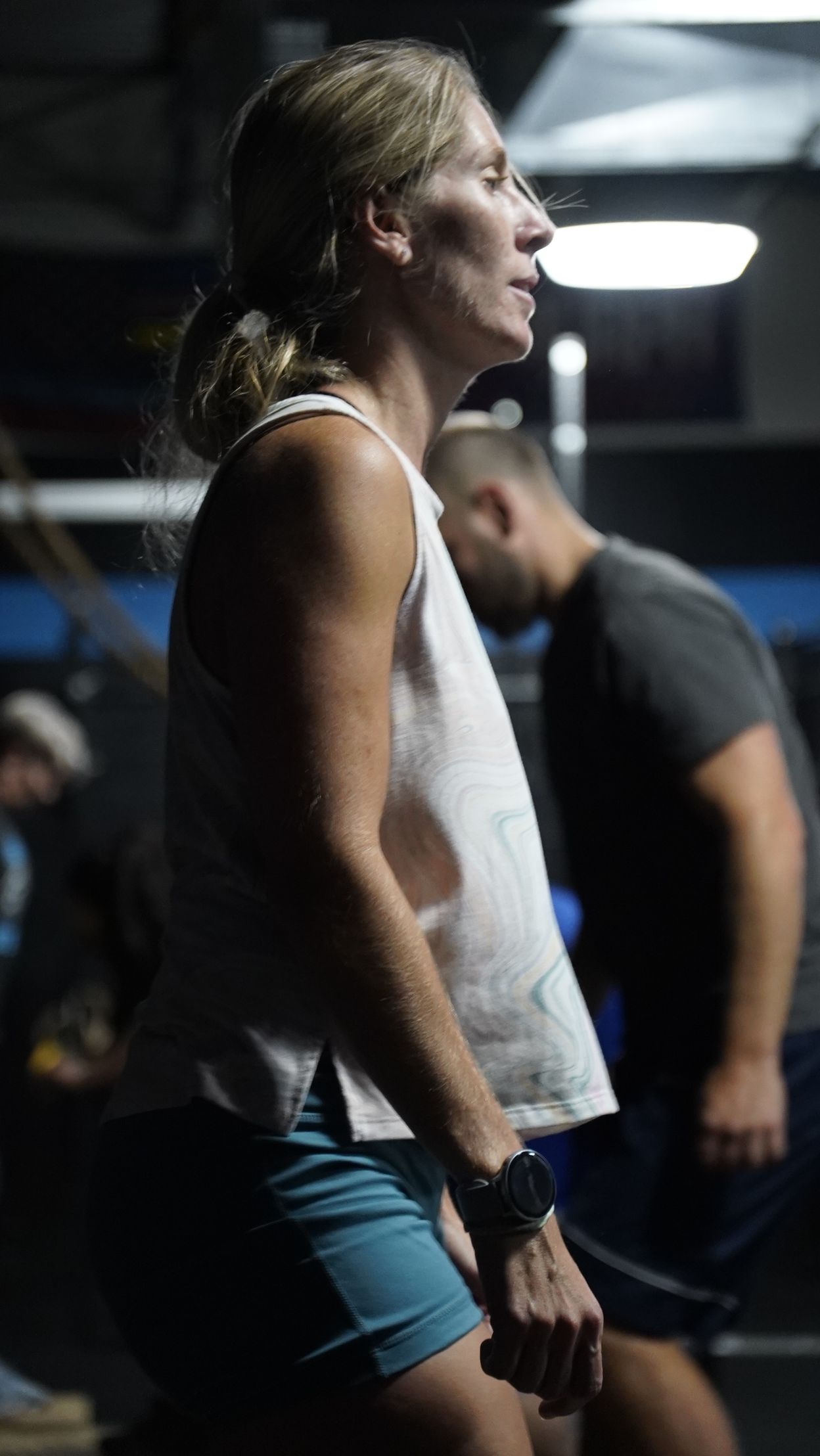 Woman in a gym, standing in profile with hands at her sides, wearing a white tank top and blue shorts.