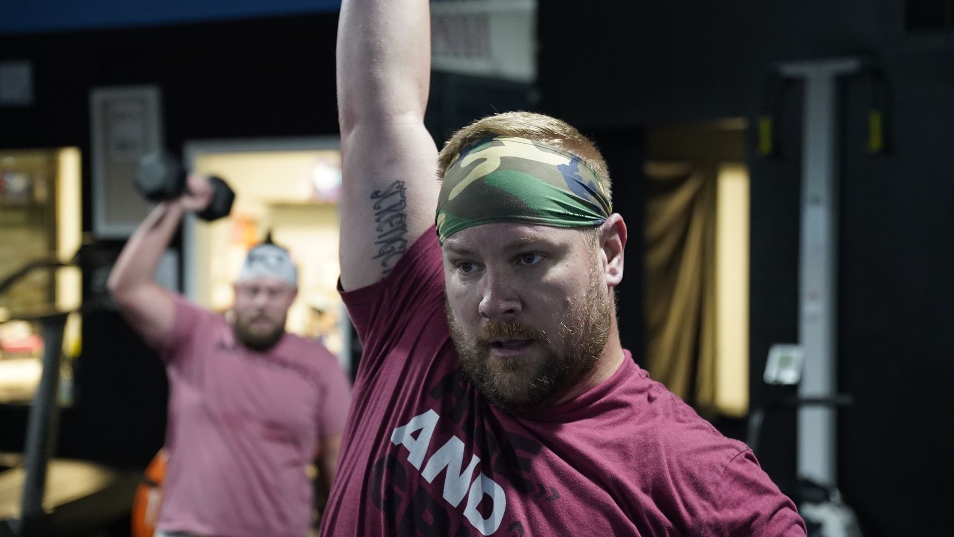 Man in maroon shirt and camo headband lifting a kettlebell in a gym, with another person behind him