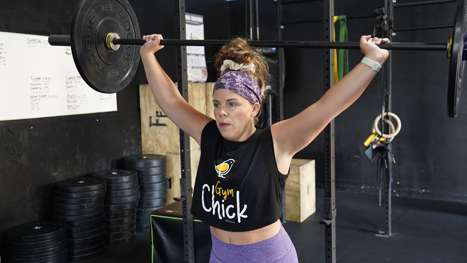 Woman lifting a barbell overhead in a gym, wearing a black shirt and purple shorts.