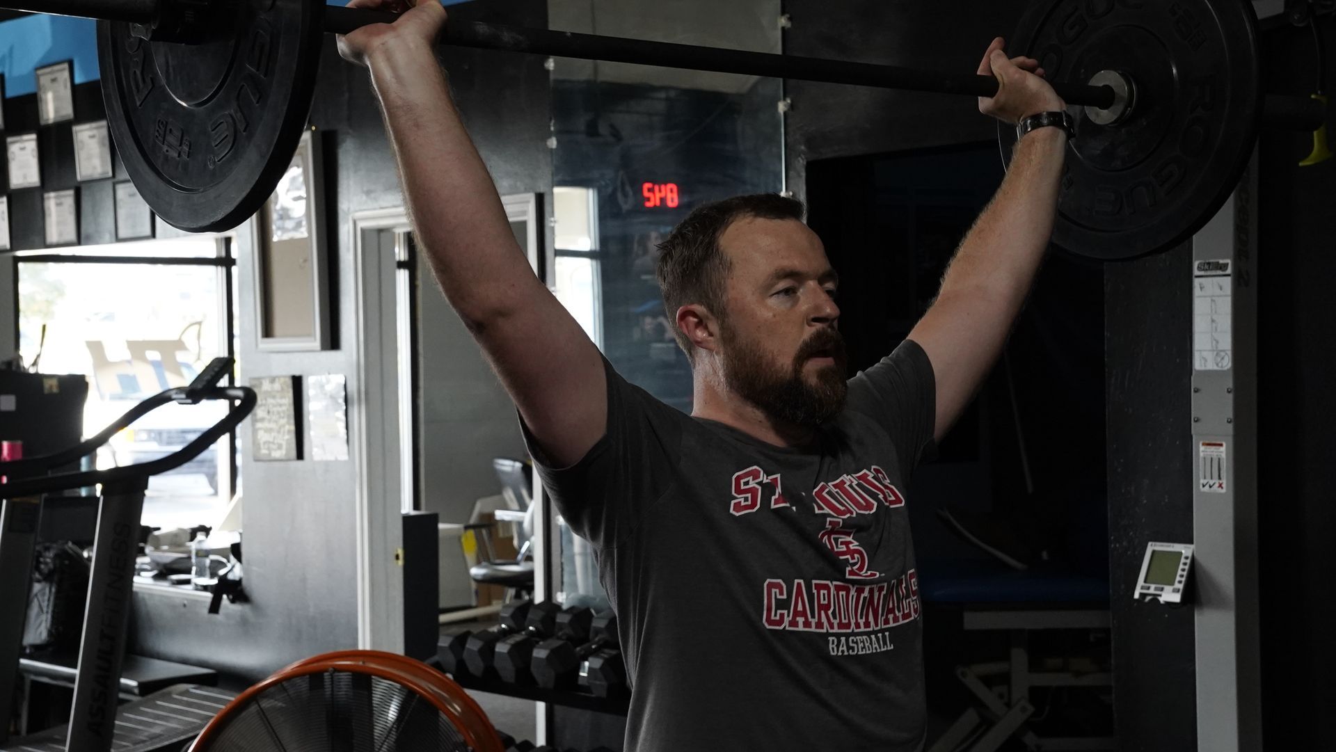 Person lifting a barbell overhead in a gym, wearing a black T-shirt and focused expression.