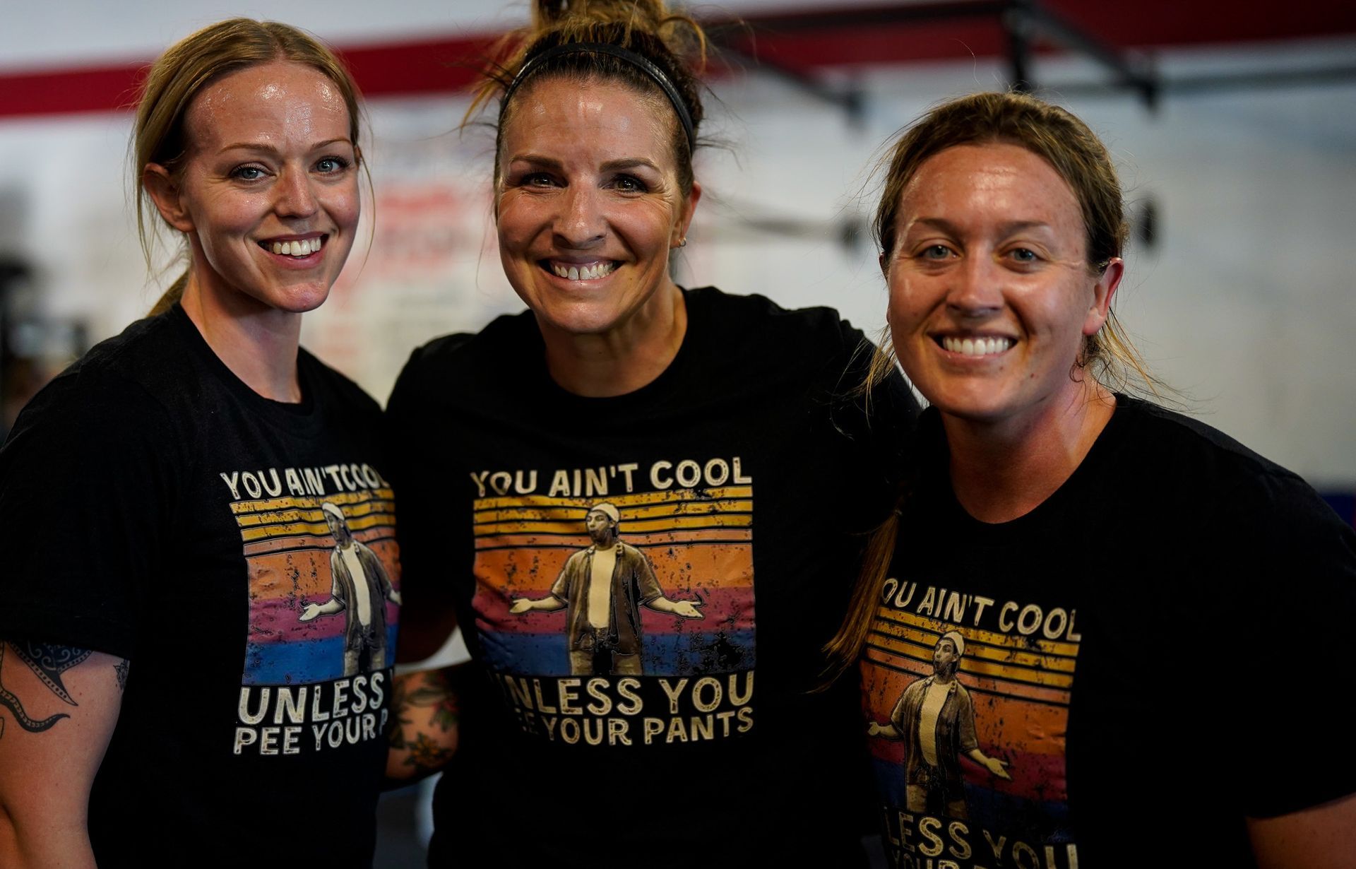 Three smiling women in black T-shirts with a colorful slogan, standing shoulder to shoulder indoors.