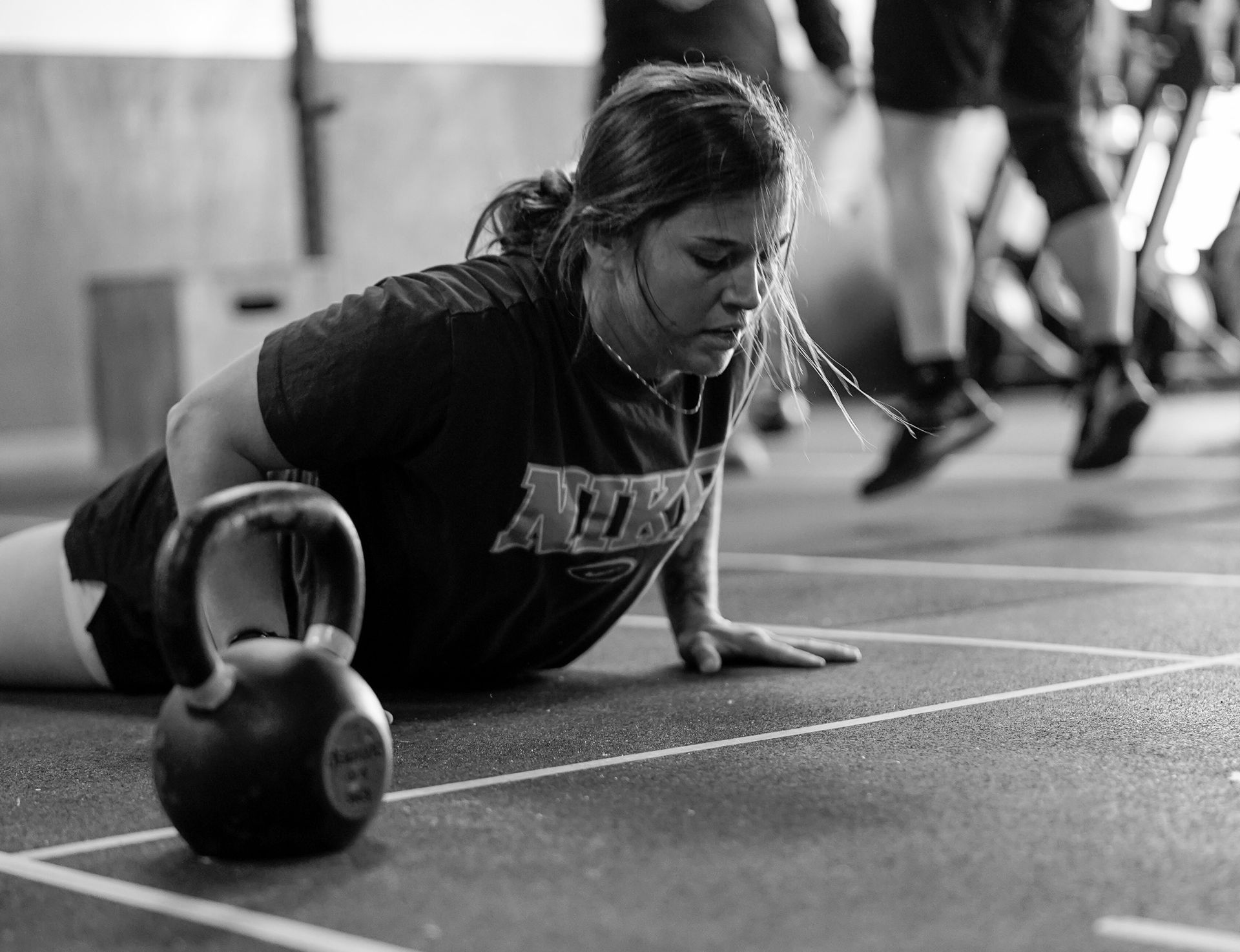 Woman doing push-ups beside a kettlebell in a gym, in black and white.