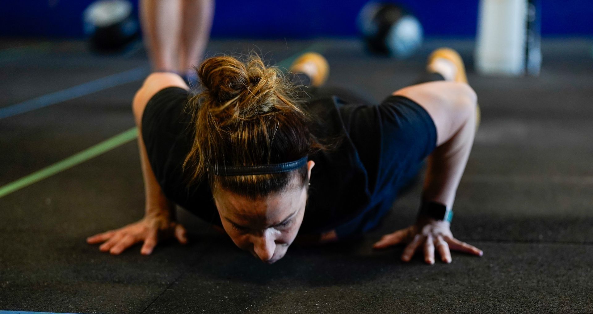 Woman doing a push-up on a gym floor, focused downward, with blurred fitness equipment in the background