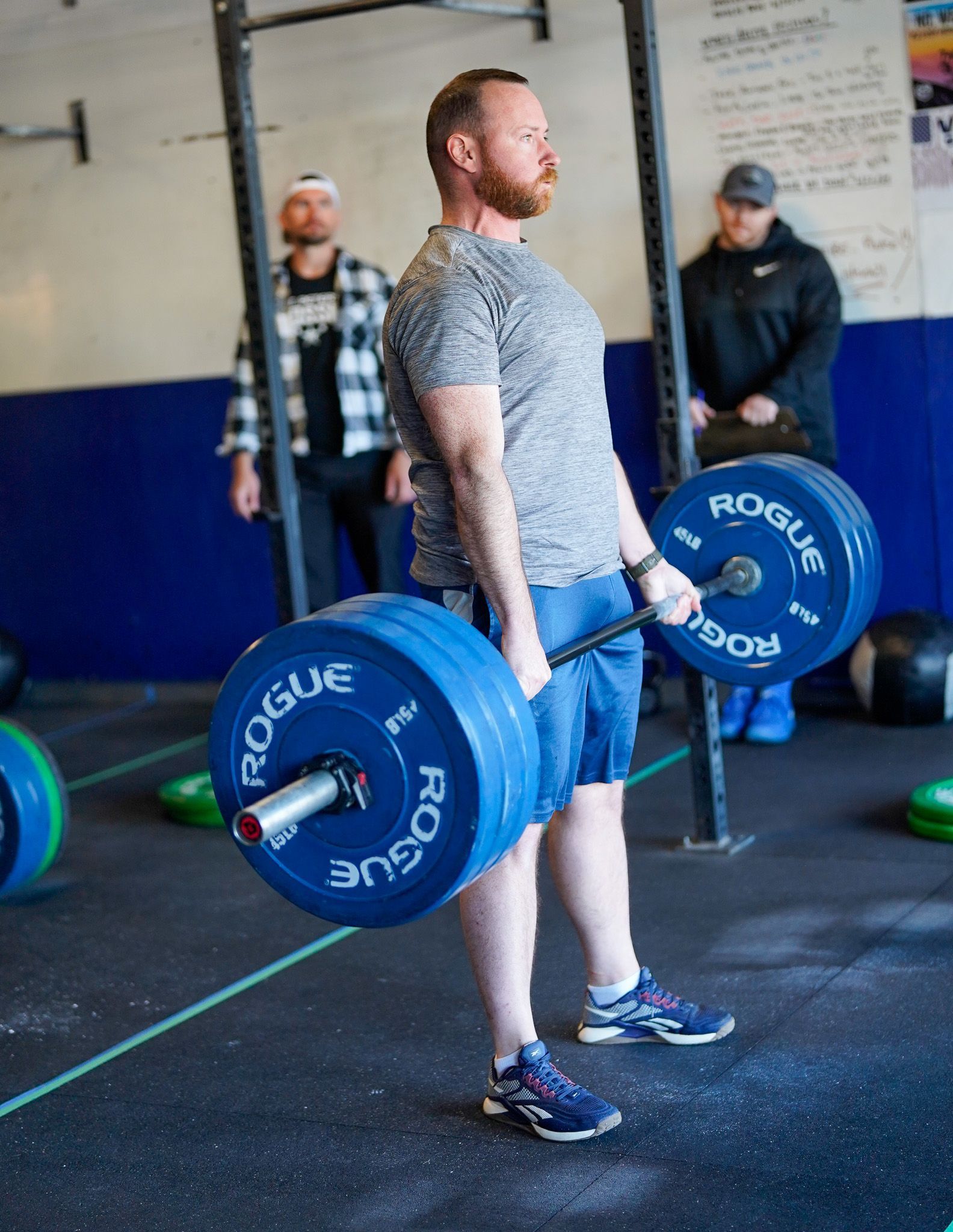 Person lifting a barbell with blue weights in a gym, standing in profile near a chalkboard.