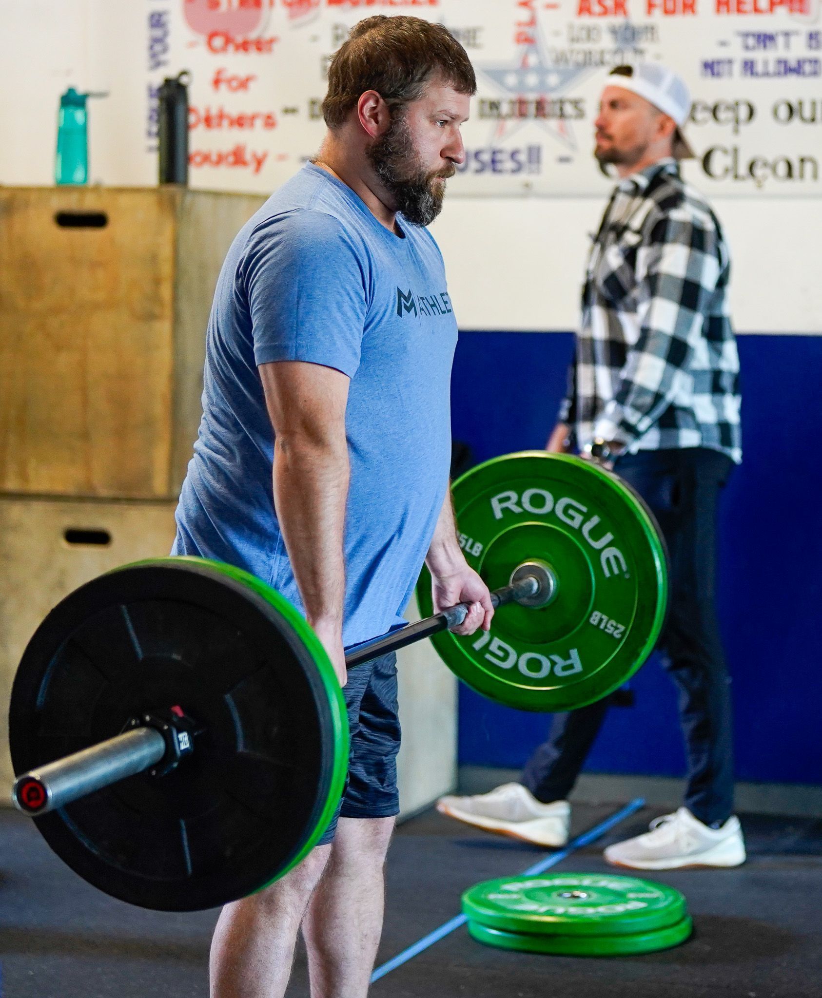 Man lifting a barbell with green plates in a gym, with another person in the background.