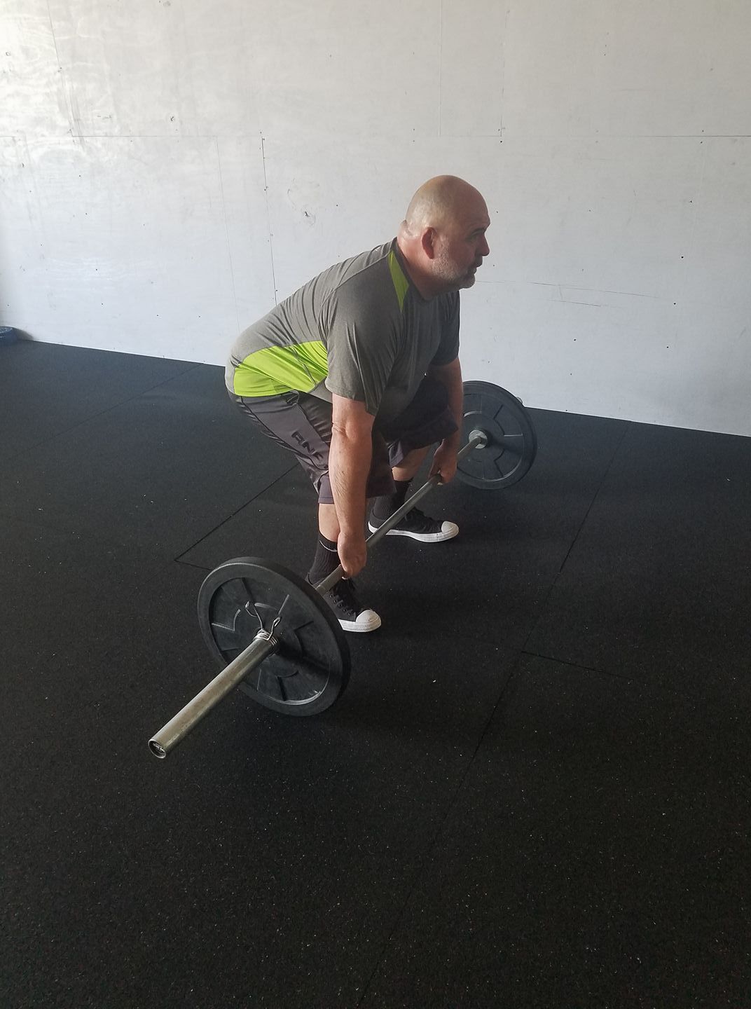 Man deadlifting a barbell in a gym on a black rubber floor, wearing a gray shirt and neon green shorts