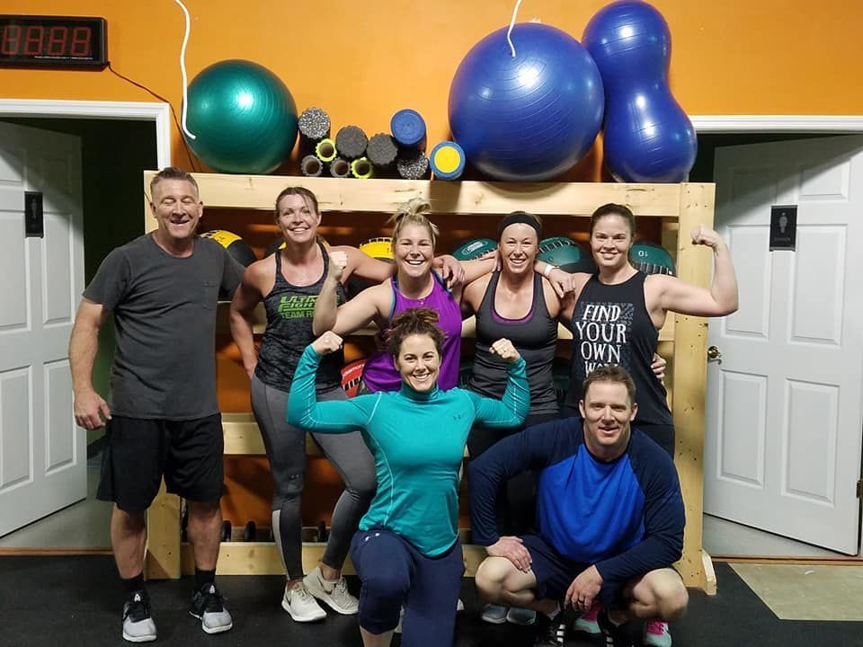 Group of people posing in a gym with exercise balls and weights on shelves, smiling in workout clothes