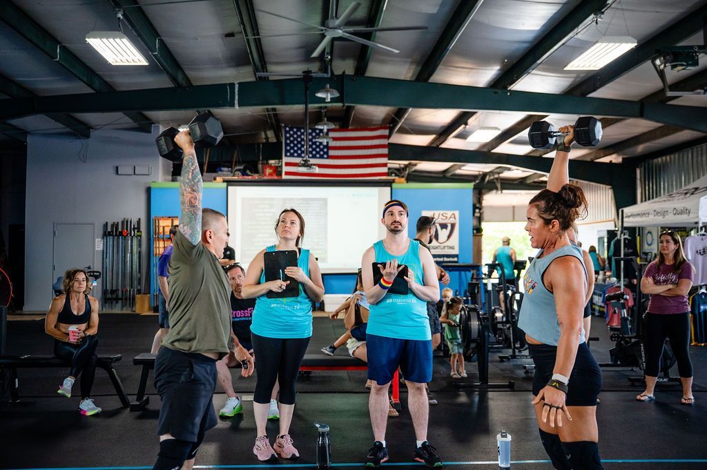 People exercising in a CrossFit gym with barbells and monitors, one person coaching in the center