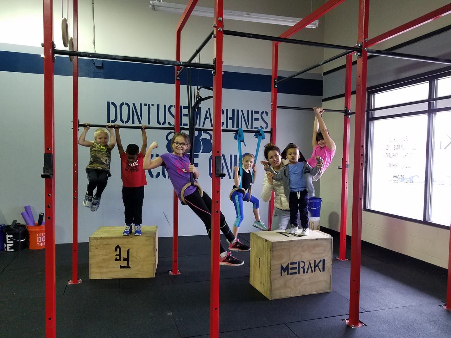 Children hanging from red gym bars in a fitness studio with wooden boxes and large windows.