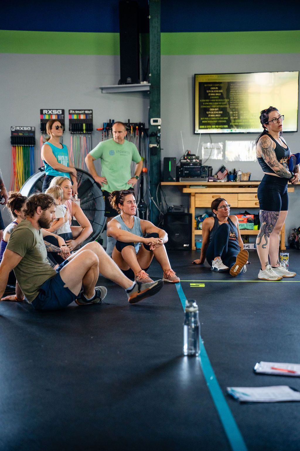 People seated on gym floor watching an instructor speaking near a counter in a bright studio.