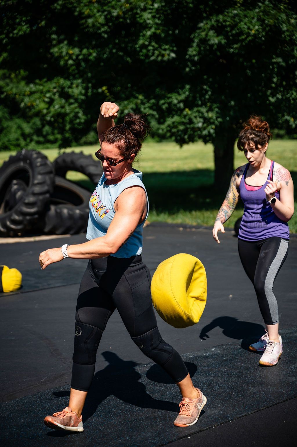 Two women exercising outdoors on a sunny day, stepping with yellow sandbags on a black mat.