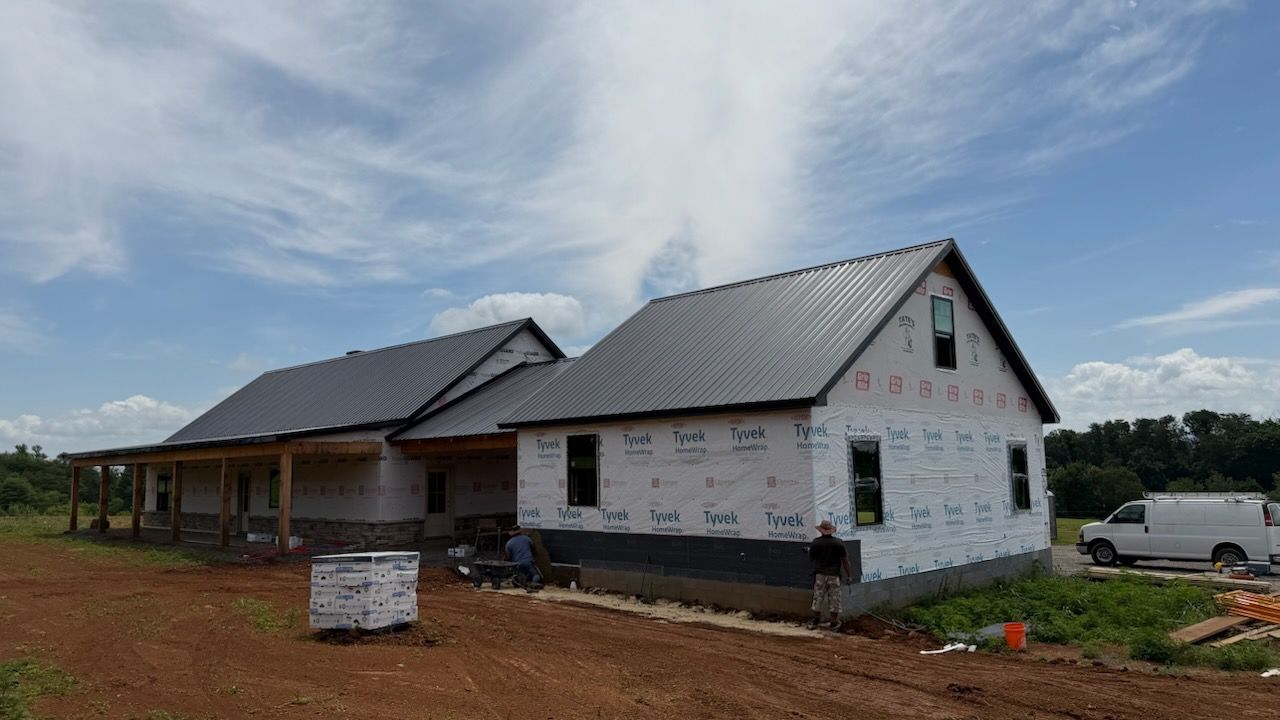 Partially built white house with black metal roof on a dirt lot under a cloudy sky