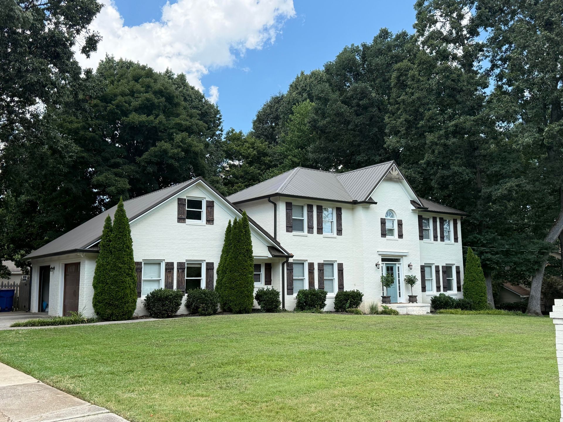 White two-story house with dark shutters, a garage, and a large green lawn under a blue sky.