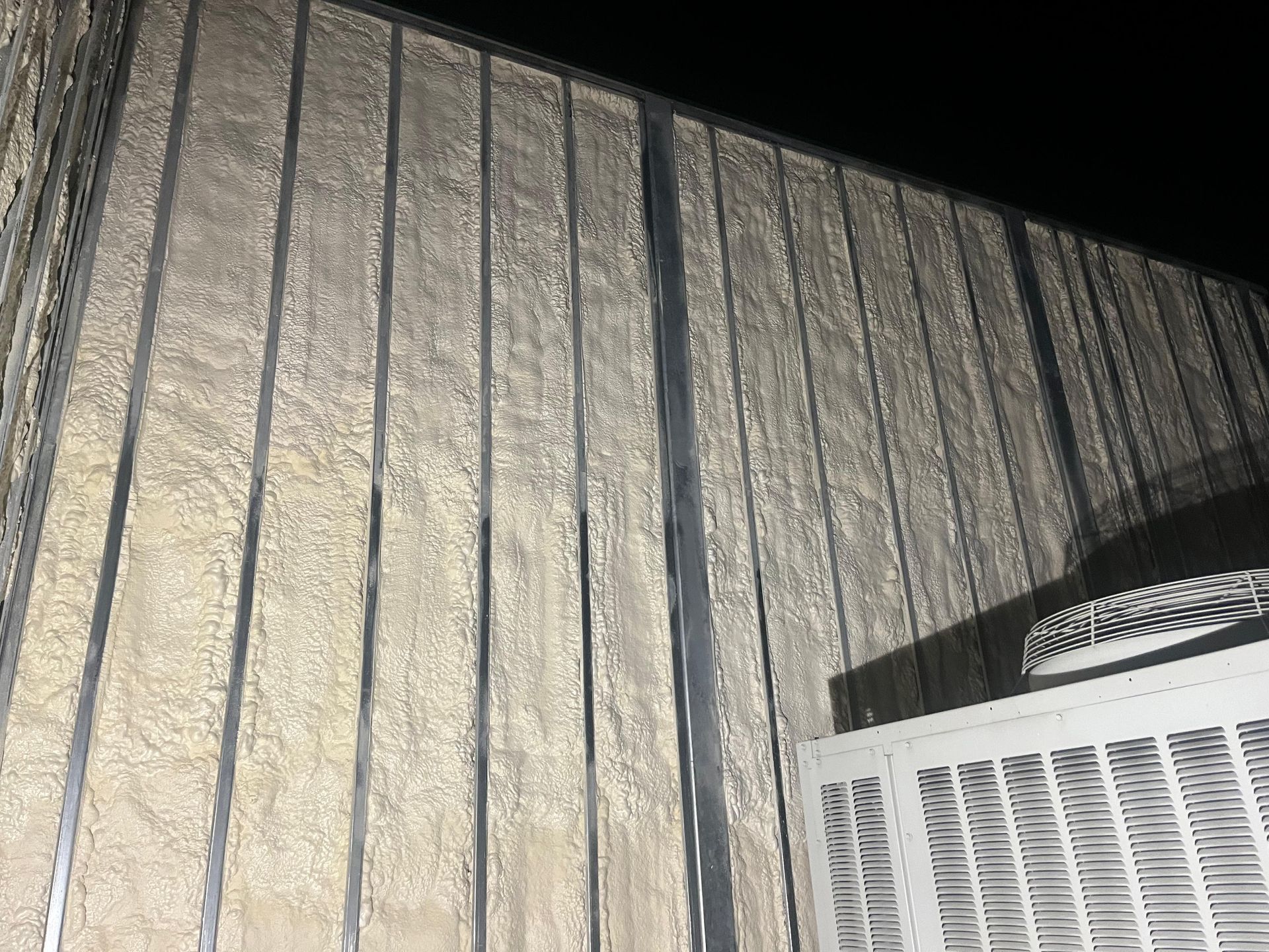 Exterior wall with vertical metal framing and a beige, textured infill. Air conditioner below.