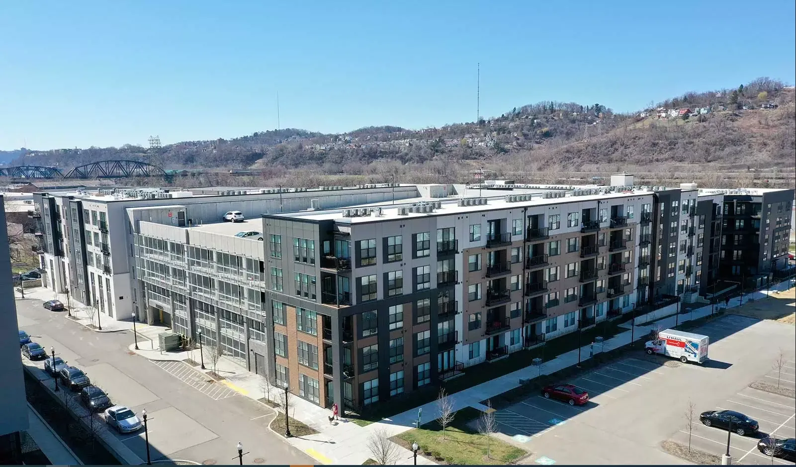 Multi-story apartment building with dark and light gray facades, parking lot, road, and hillside in background.