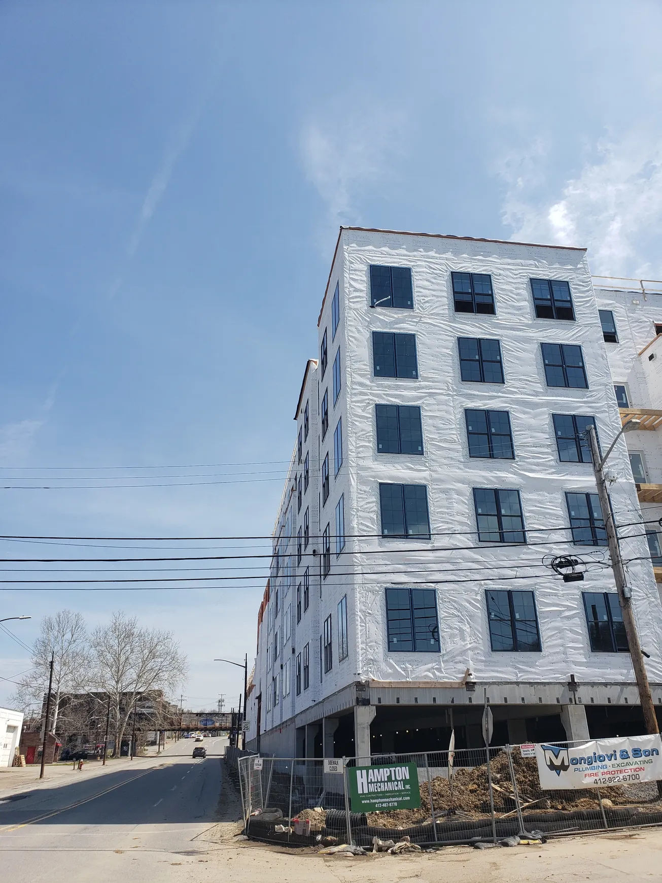 Construction of a tall, white building with dark windows under a blue sky. Wires and a street in the foreground.