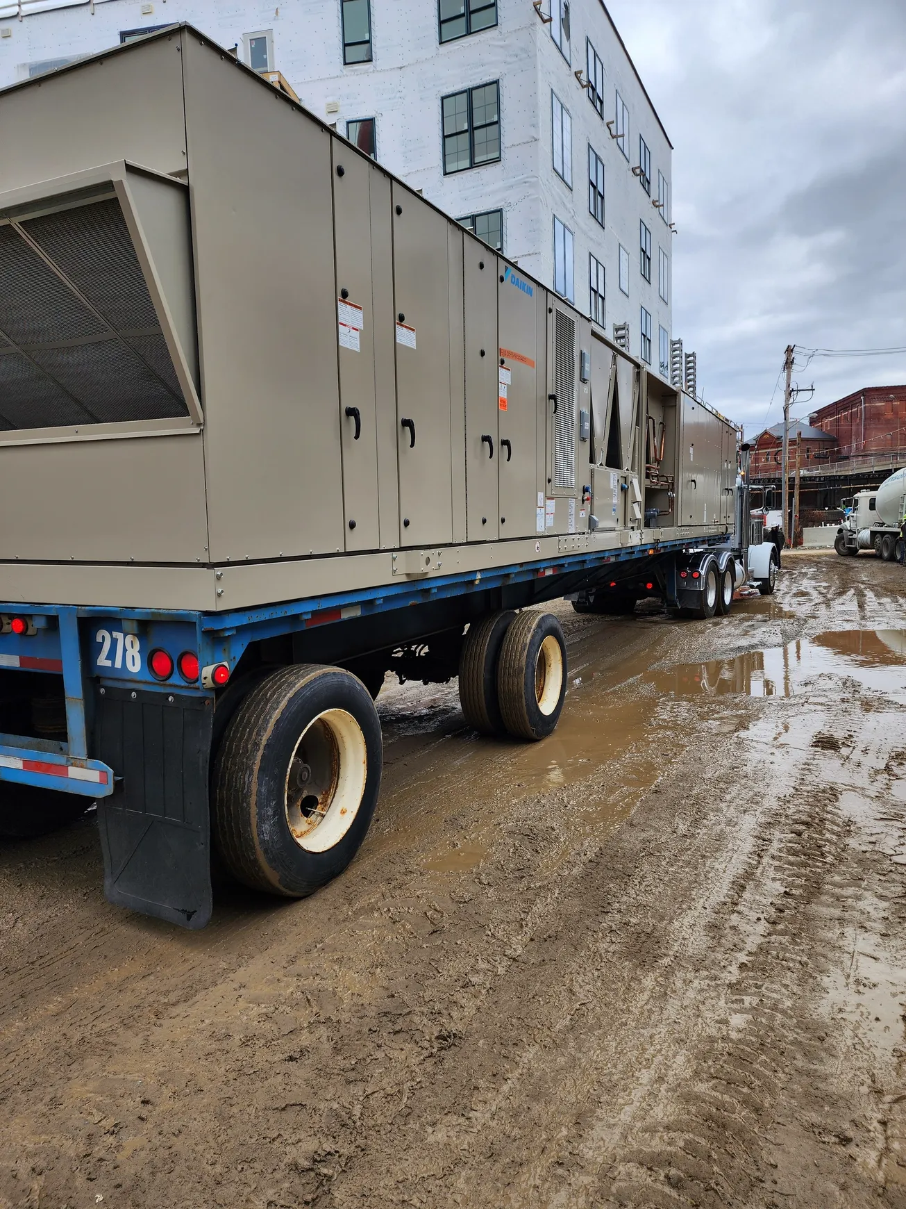 Large HVAC unit on a flatbed trailer at a construction site. Muddy ground, building in background.