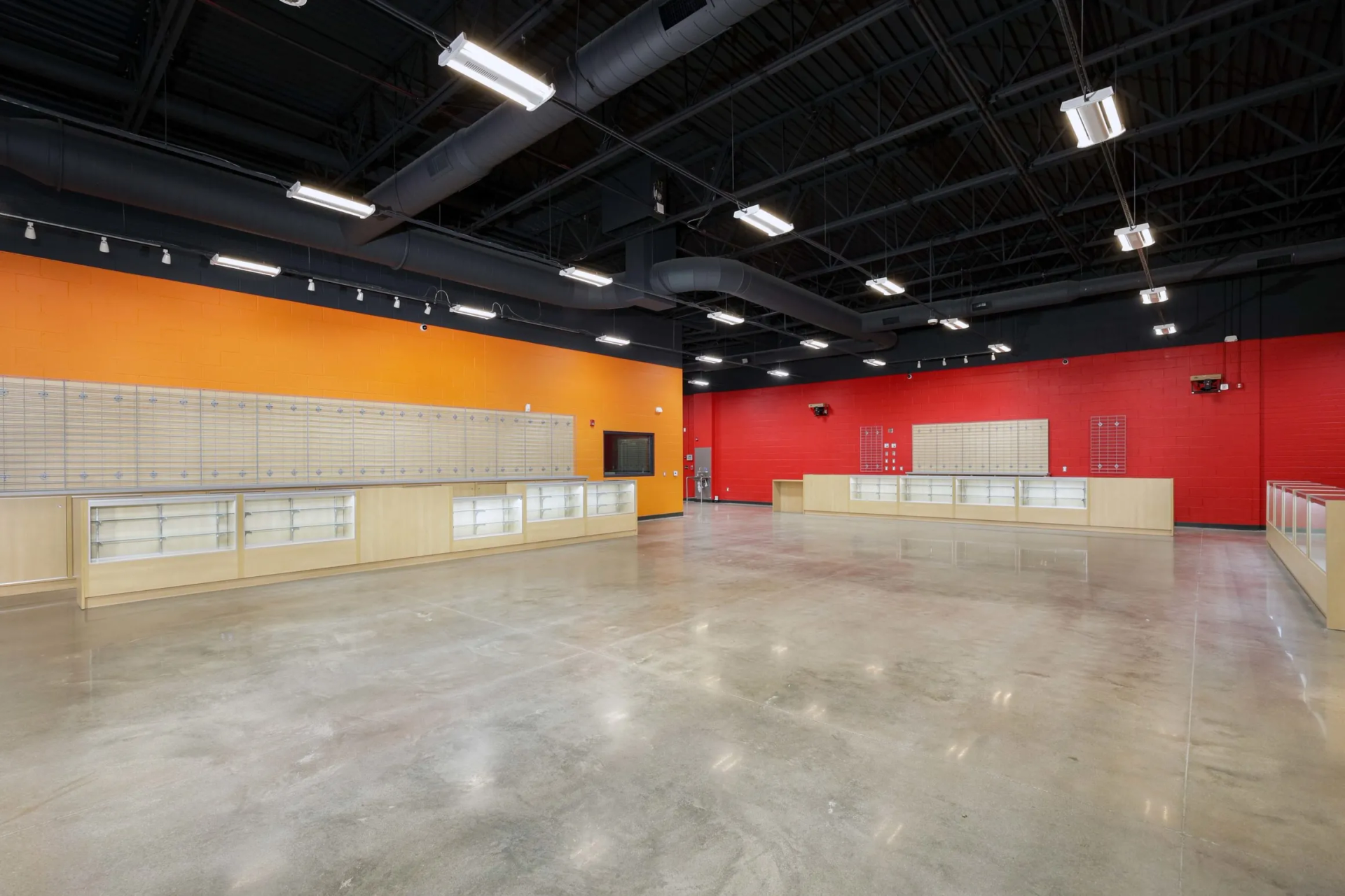 Empty retail space with orange and red walls, concrete floor, and display counters. Black ceiling with lights.