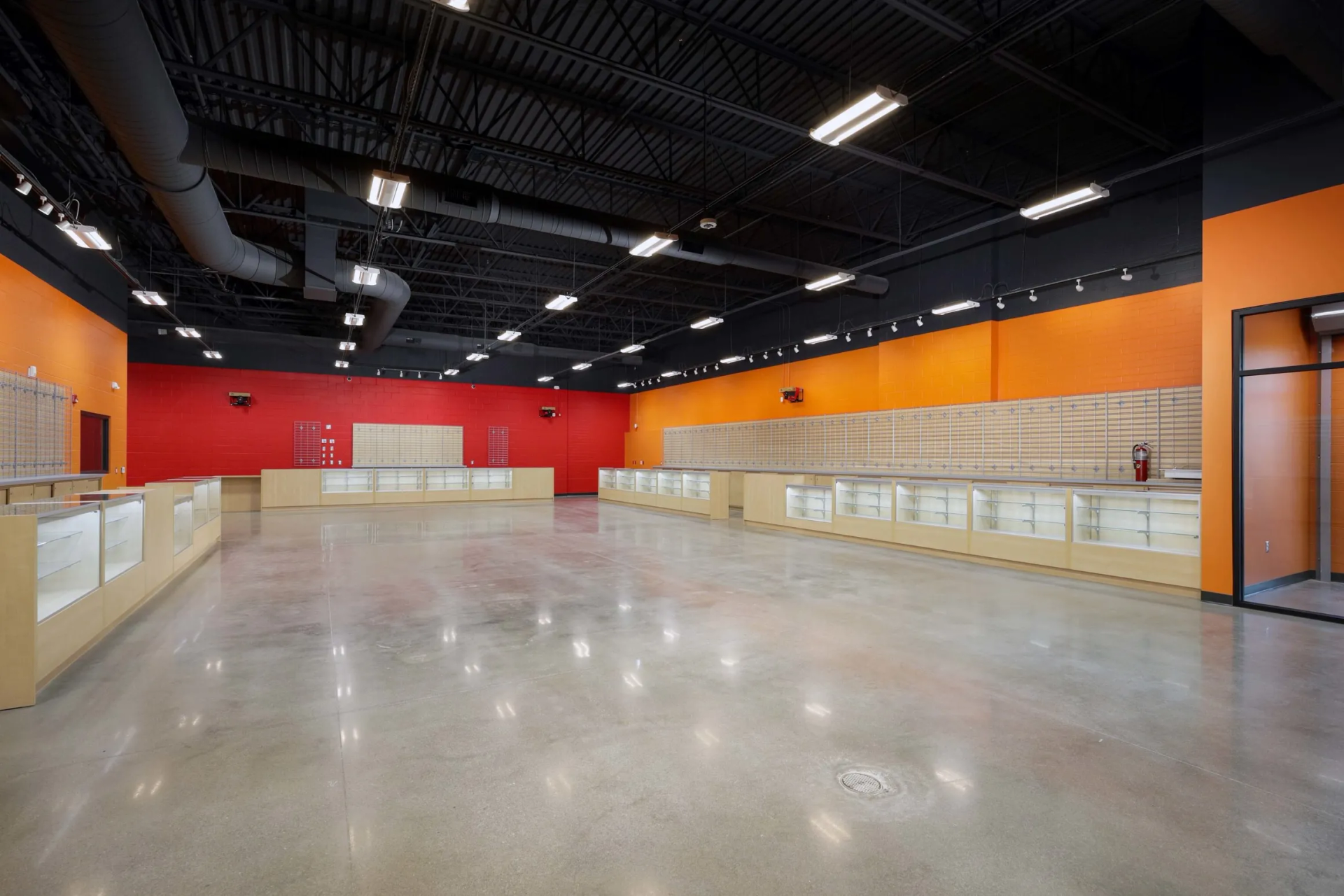 Empty retail space with polished concrete floor, light wood displays, and bright orange and red walls.