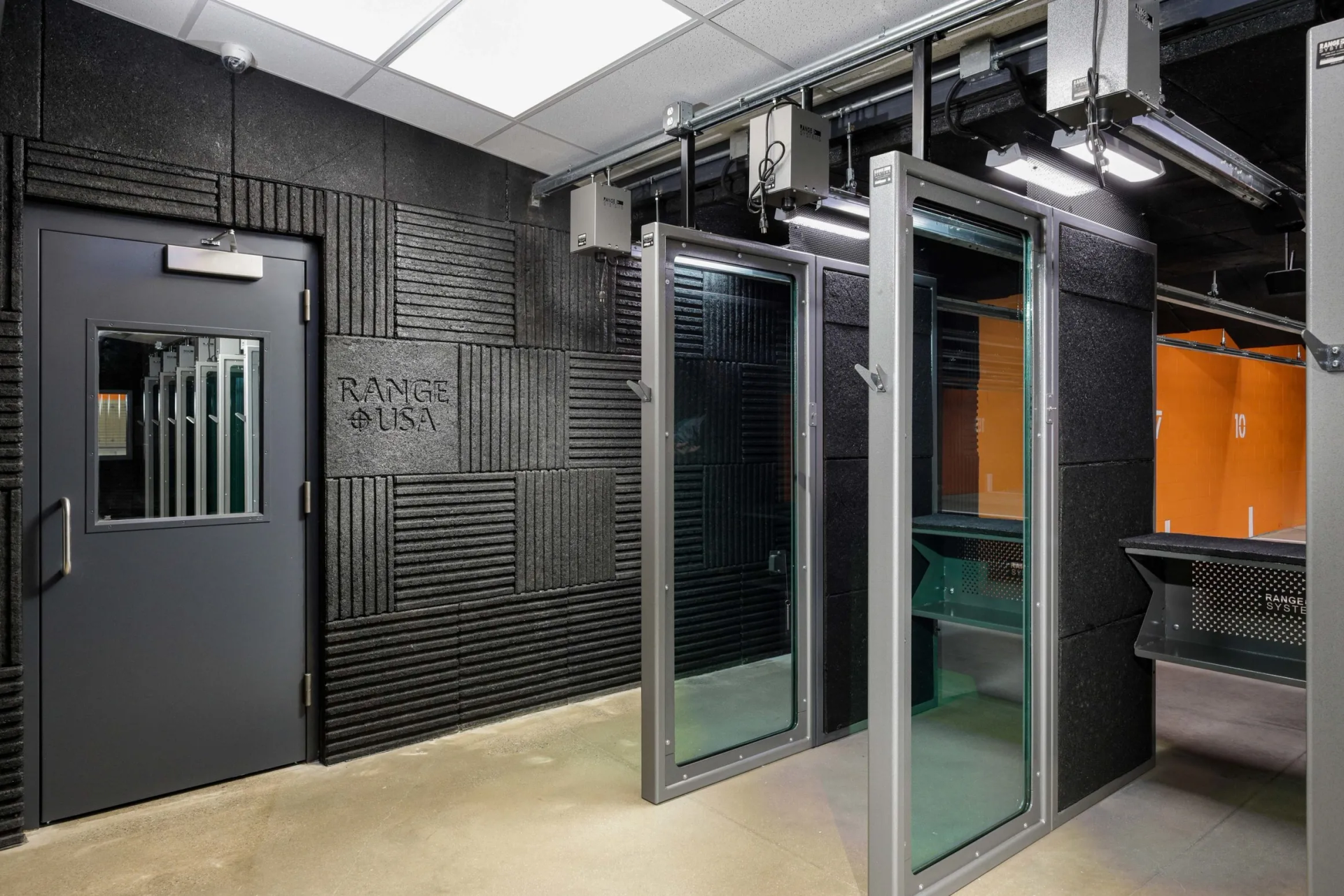 Shooting range interior: soundproofed walls, booths with glass, metal door, and ceiling lighting.