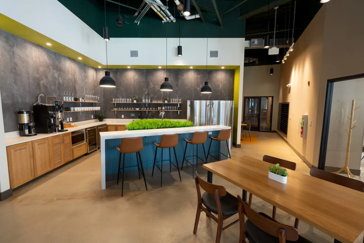 Modern office breakroom with a kitchen, bar, and dining table; teal, wood, and concrete accents.