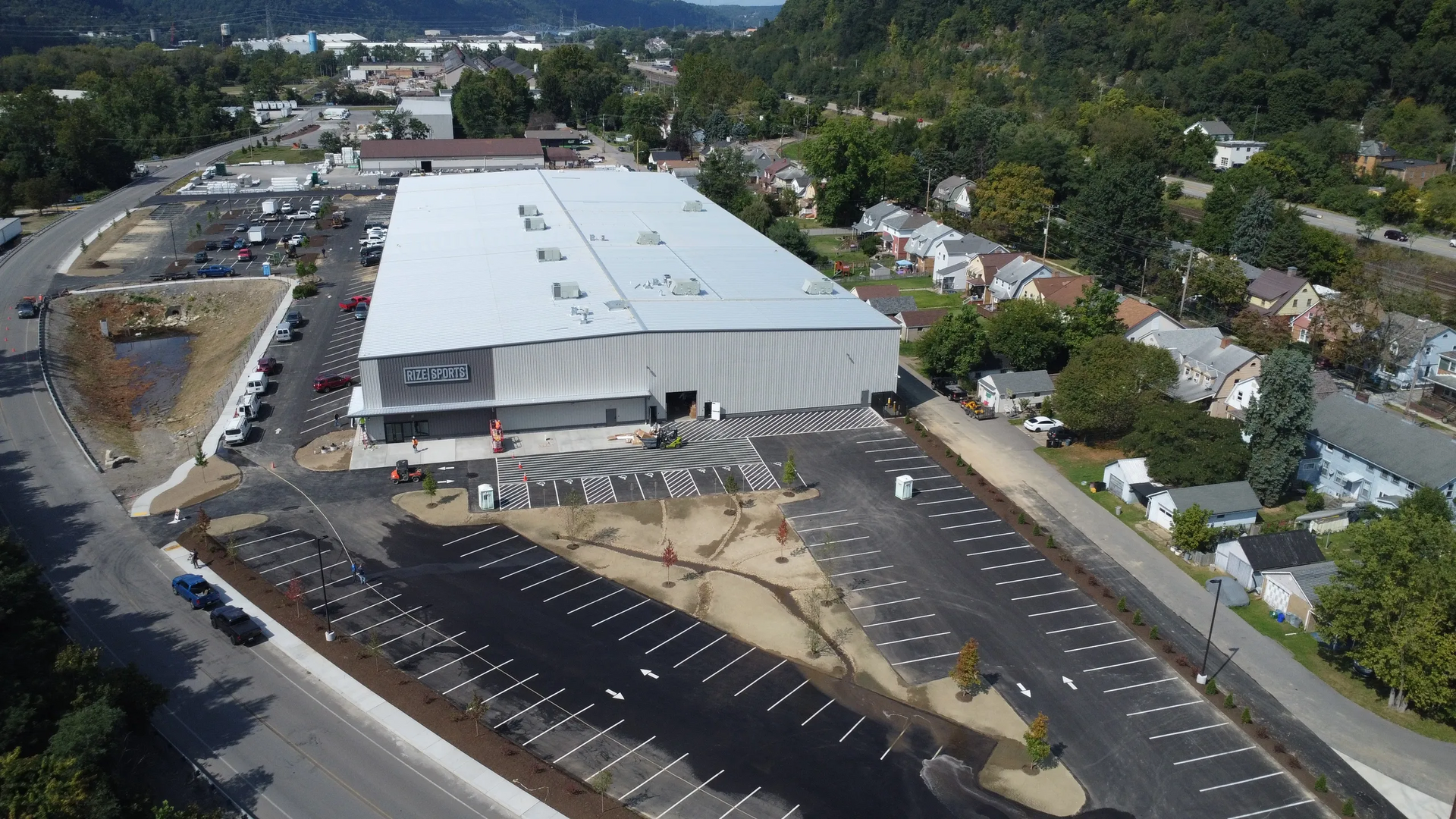Aerial view of a large commercial building with extensive parking, a town and hills in the background.