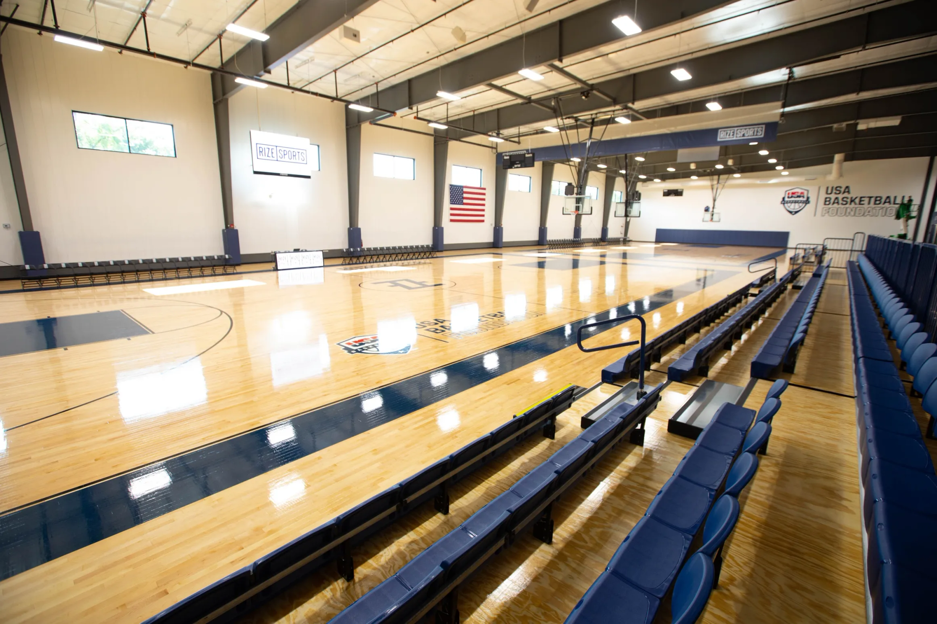 Basketball court with bleacher seating, American flag, and indoor lighting.