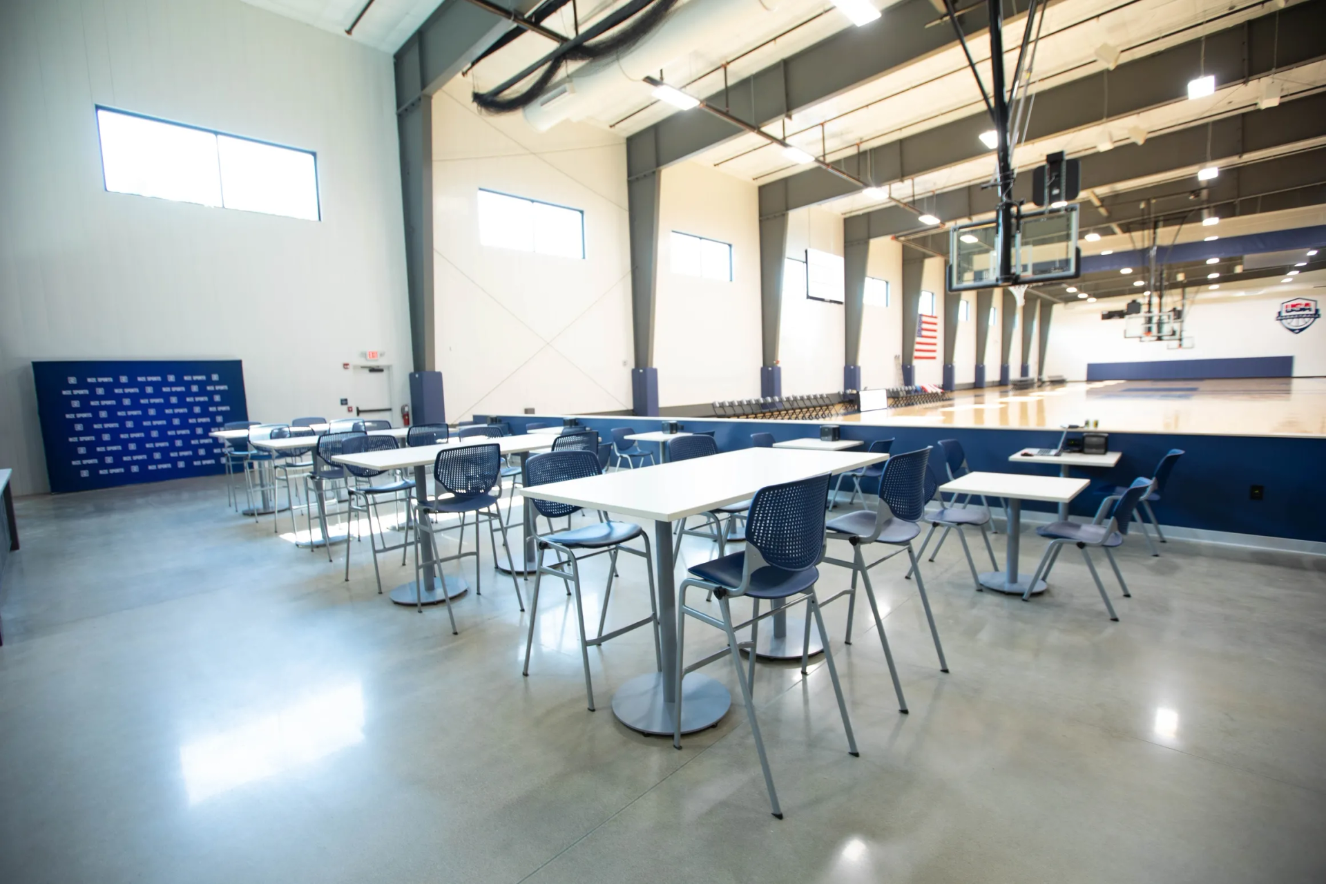 Tables and chairs set up in front of a gymnasium, blue and gray color scheme, basketball hoops visible.