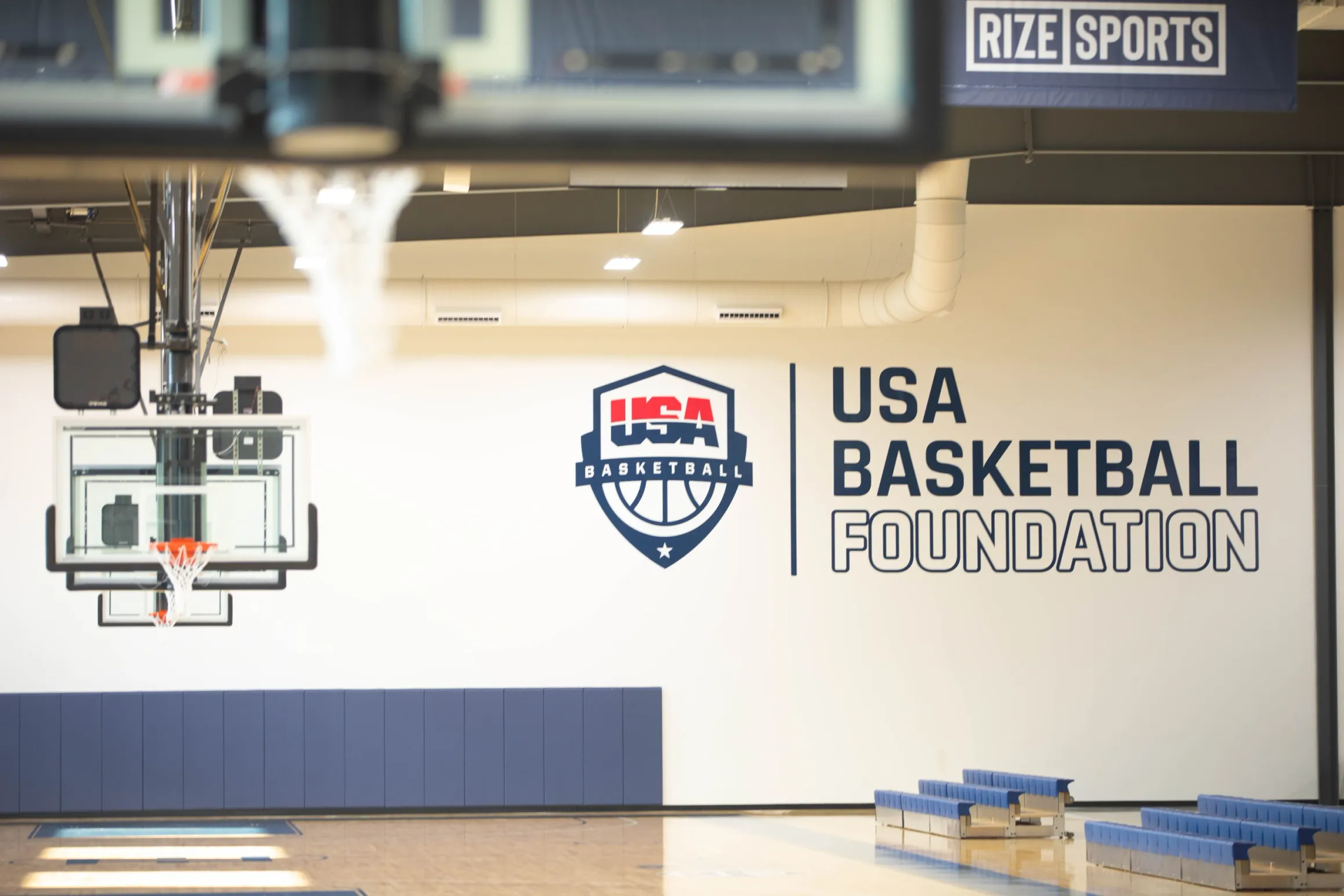 Basketball court with USA Basketball Foundation logo on the wall. Hoop and bleachers visible.