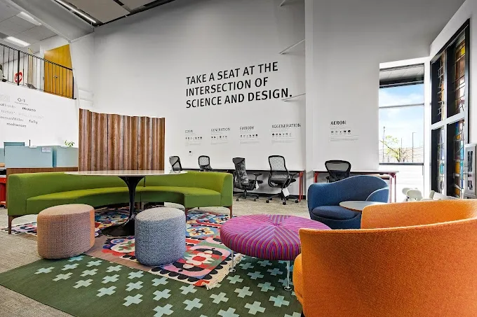 Colorful office lounge with sofas, chairs, ottomans, and a rug. Text on the wall: Take a seat at the intersection of science and design.