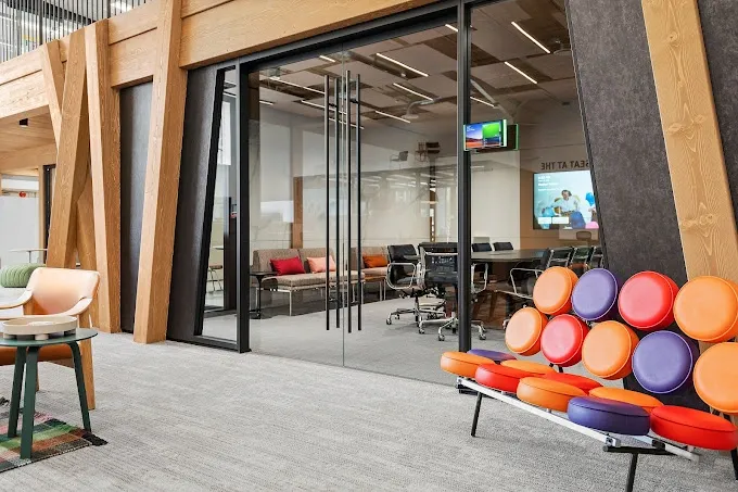 Modern office space with glass-walled meeting room, colorful seating, and wood accents.