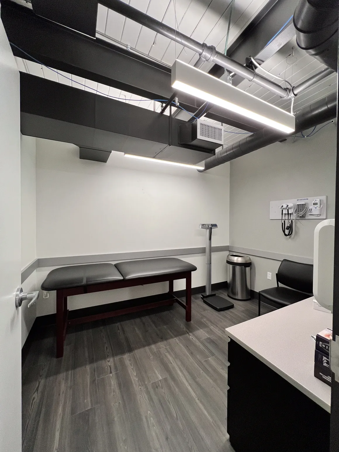 Examination room with an exam table, scale, chair, and blood pressure monitor. Gray walls and flooring.