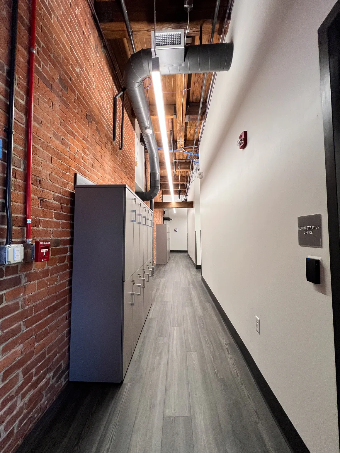 Long, narrow hallway with brick wall and lockers on one side, white wall on the other.