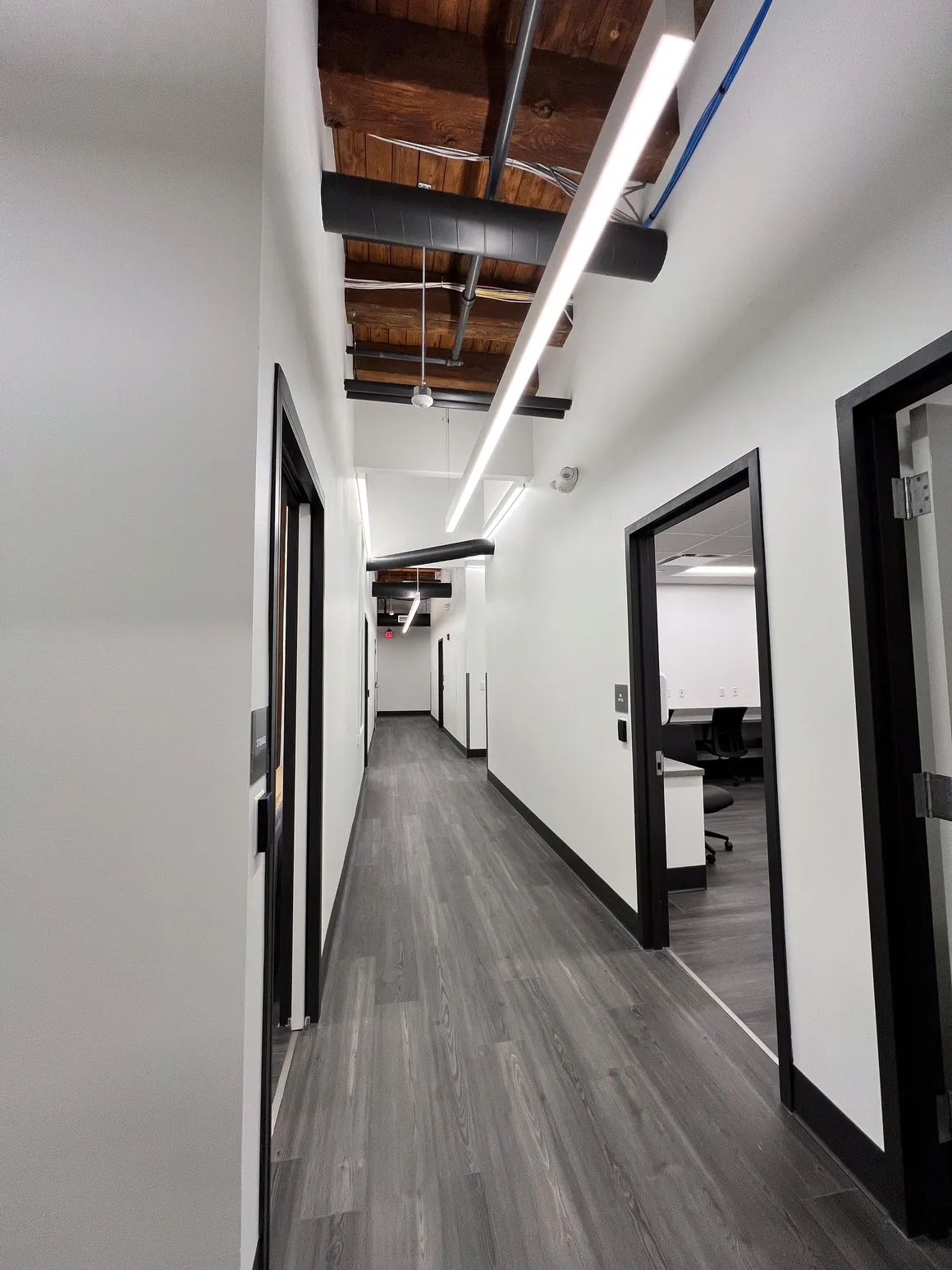 Narrow office hallway with white walls, dark gray floor, black door frames, and bright overhead lights.
