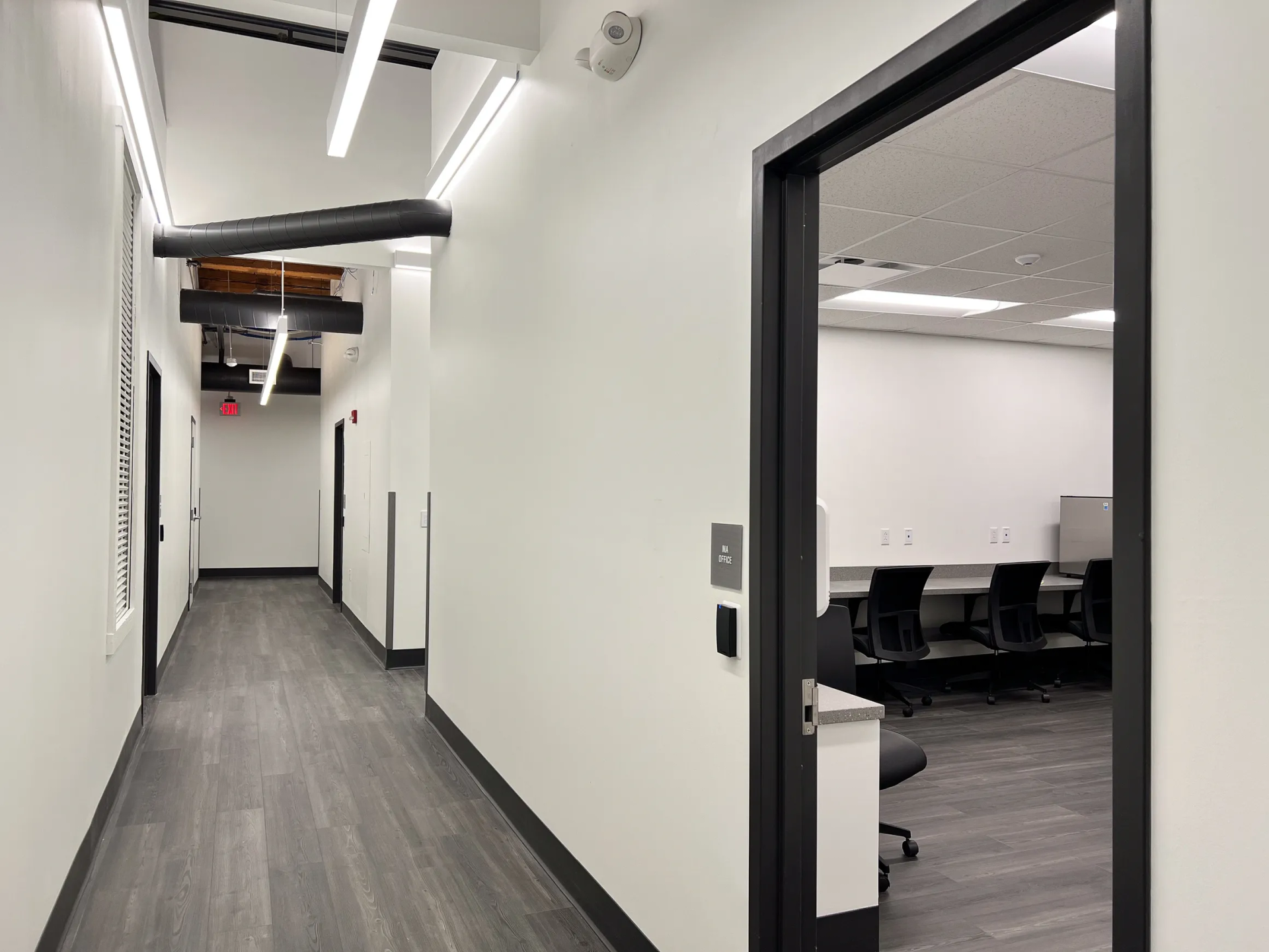 Hallway with white walls, dark gray floor, open door reveals a room with chairs and tables.