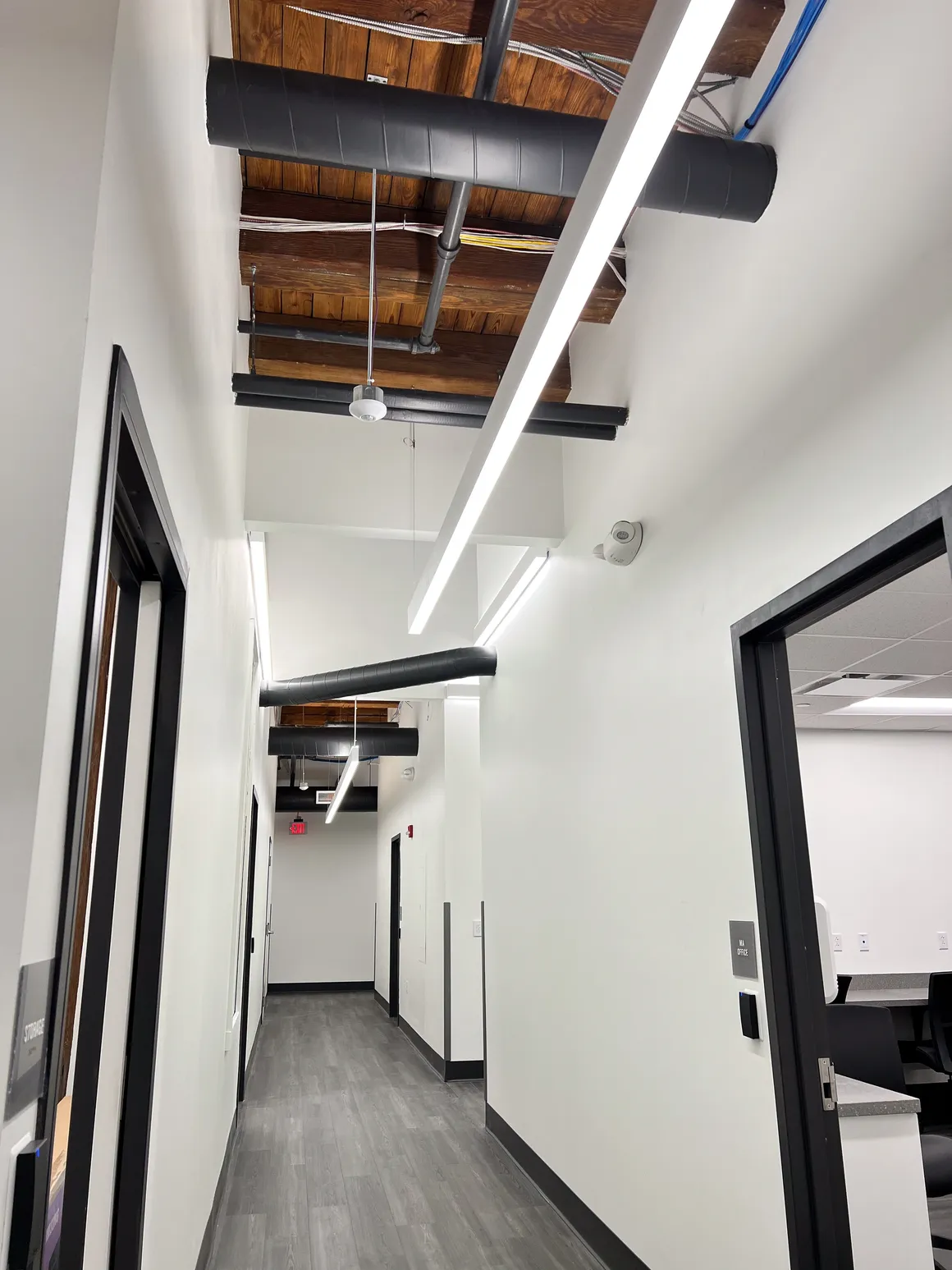 Narrow office hallway with white walls, gray floor, black door frames, and exposed ceiling with ductwork.