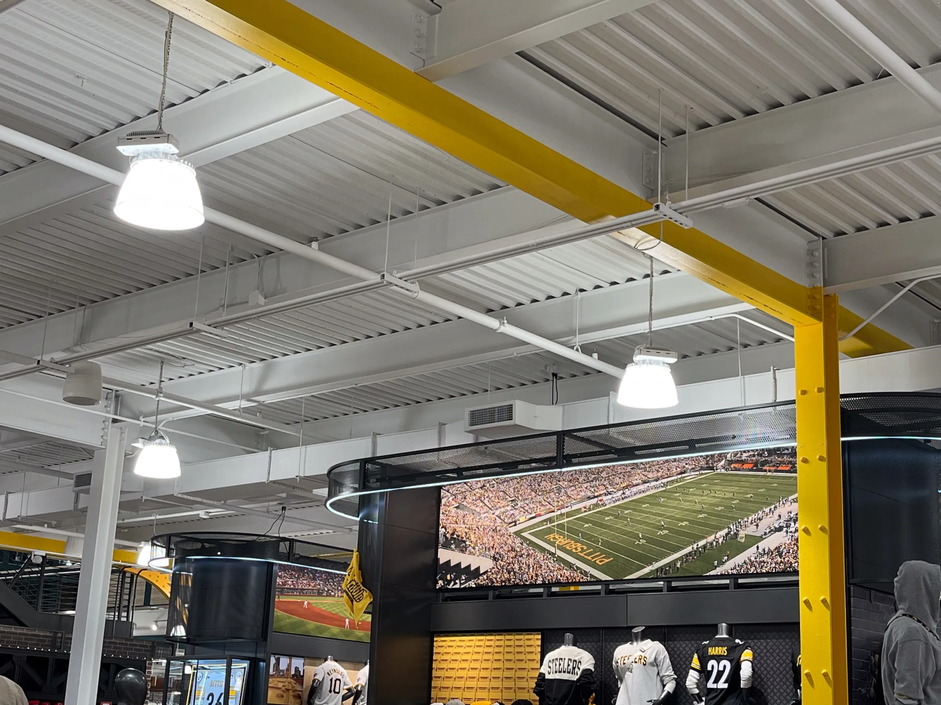 Inside a store with white ceiling, yellow beams, lights, and large TV screen displaying a football field.