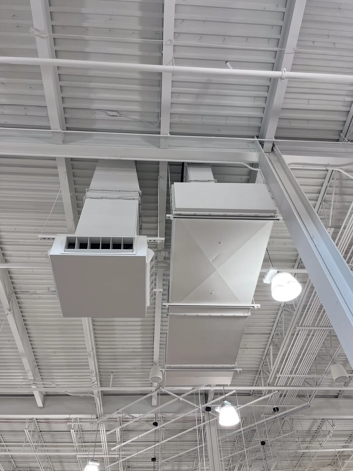 Overhead view of white HVAC ductwork in a commercial building. Metal beams and lights are also visible.