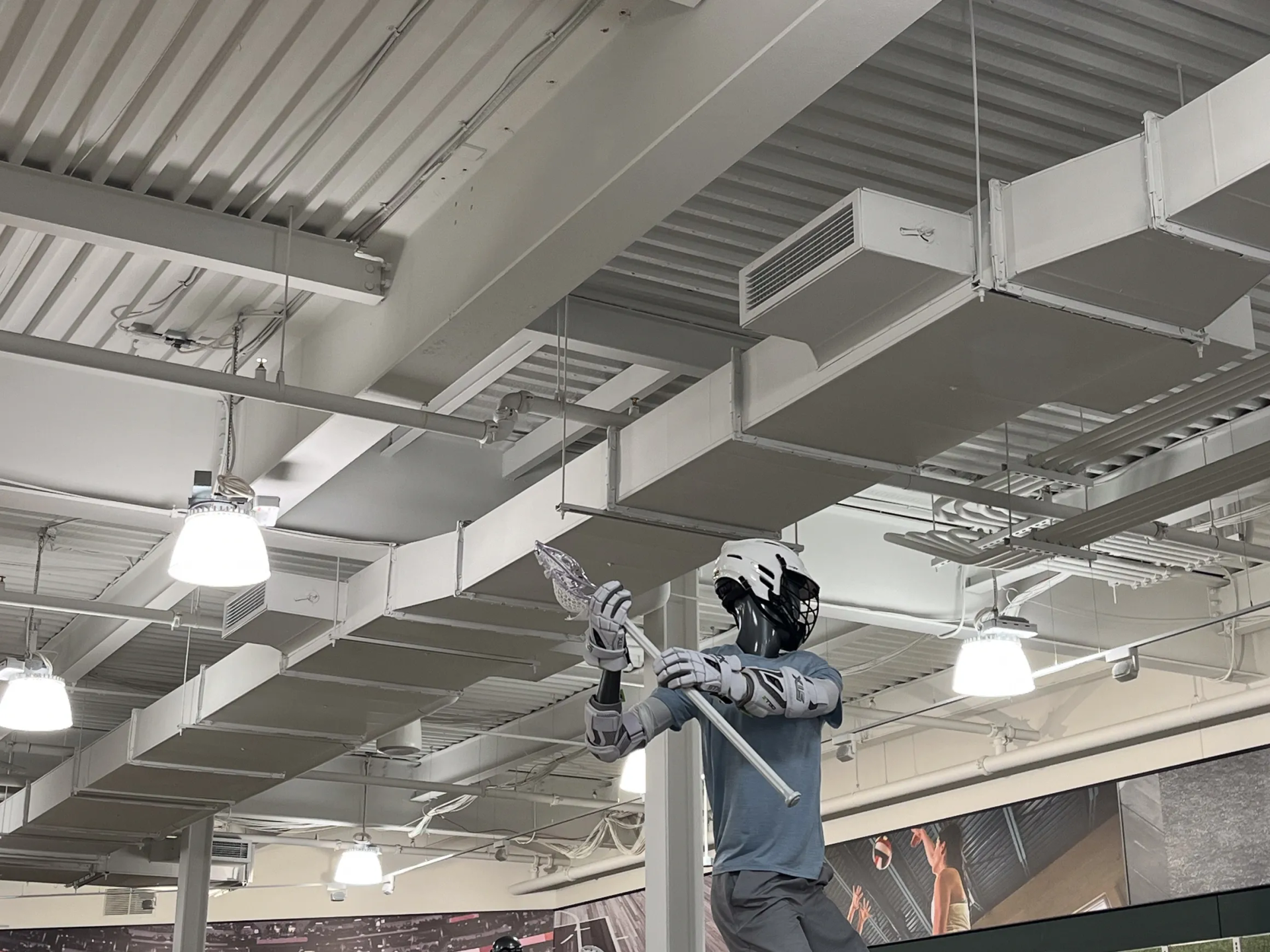 Mannequin with lacrosse stick indoors, gray and white clothing, store ceiling.