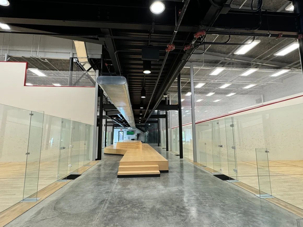 Interior view of squash courts with glass walls, a long wooden walkway, and overhead lighting.