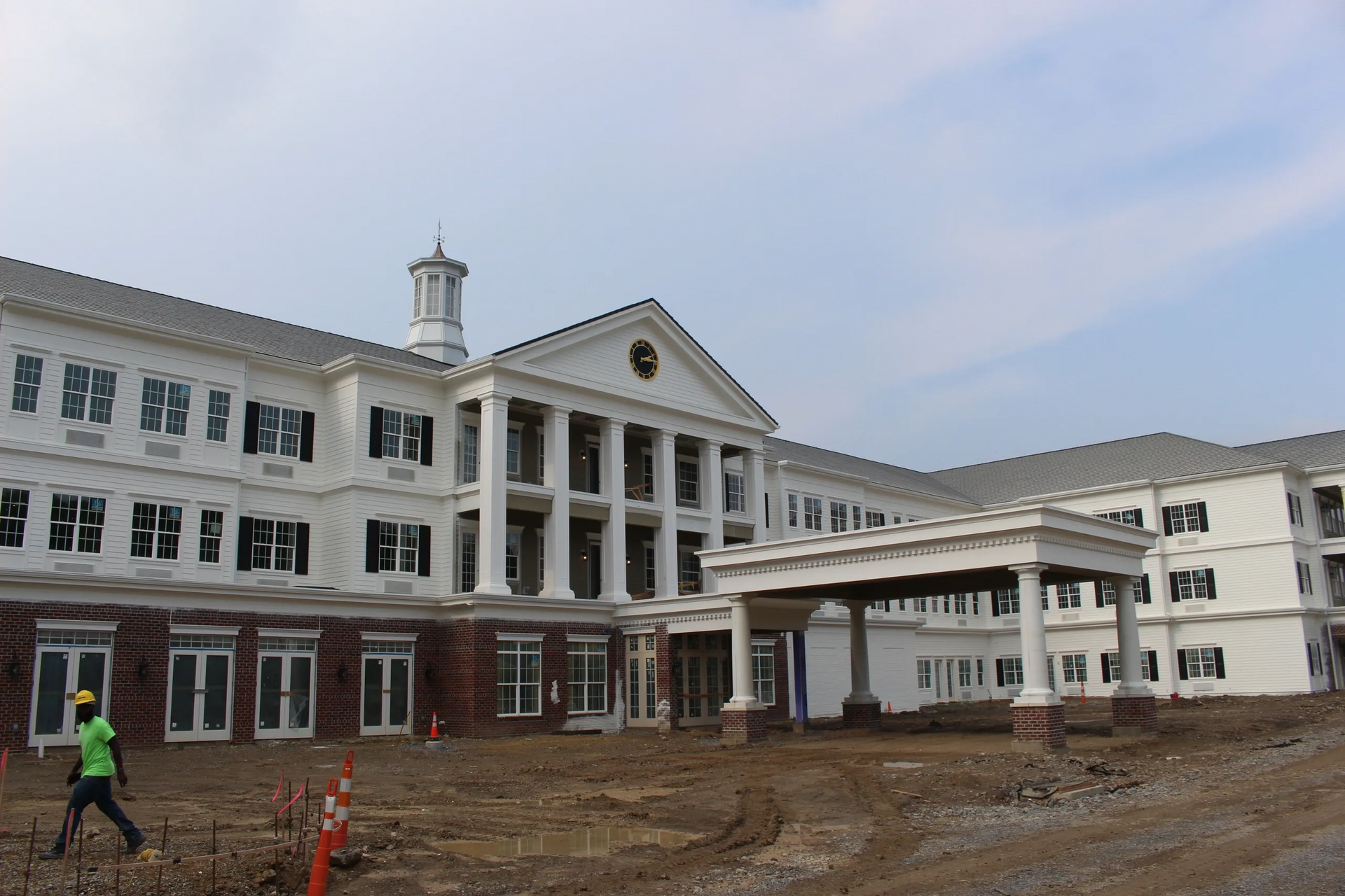 White building with columns, under construction, dirt in front, worker walking.