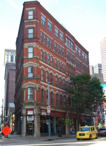 Red brick, multi-story building on a city street corner, with storefronts below. Yellow cab visible.